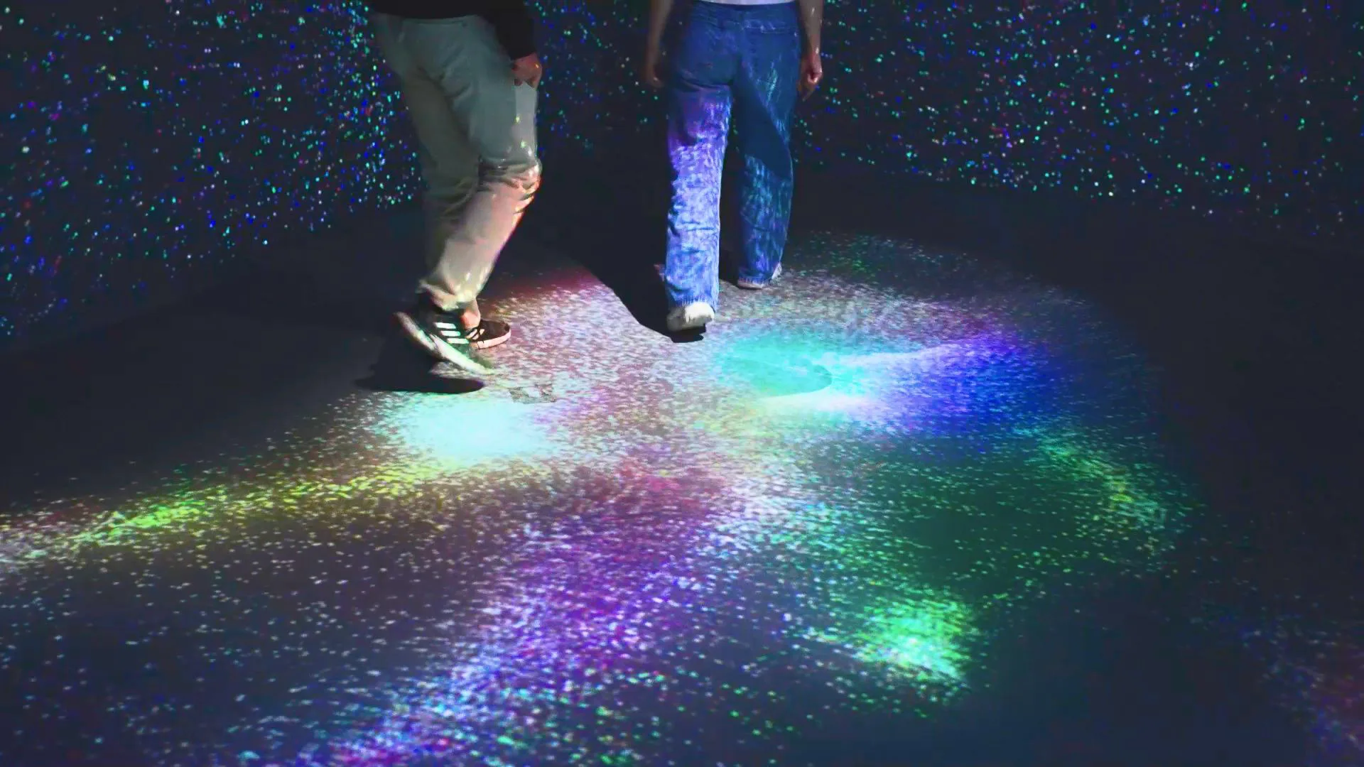 Two visitors' legs on a floor of rainbow-coloured particles pooling around their feet